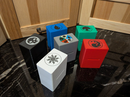 standard_mtg,standard_magic_the_gathering|Six Magic: The Gathering Standard deck boxes with mana symbol lids: black Swamp skull, blue Island drop, green Forest tree, red Mountain flame, white Plains sun and a gray five-color box
